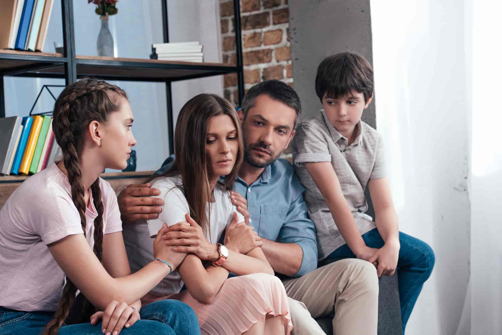 A family of four sitting close together on a couch, comforting one another during a serious conversation.